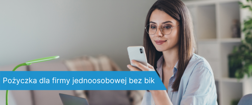 Młoda kobieta w okularach siedząca przy biurku, patrzy na telefon z uśmiechem; obok laptop i lampka